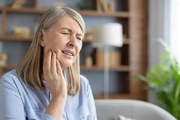 Elderly woman sitting at home, holding her jaw and grimacing in pain, indicating a dental emergency. Elderly woman sitting at home, holding her jaw and grimacing in pain, indicating a dental emergency.
