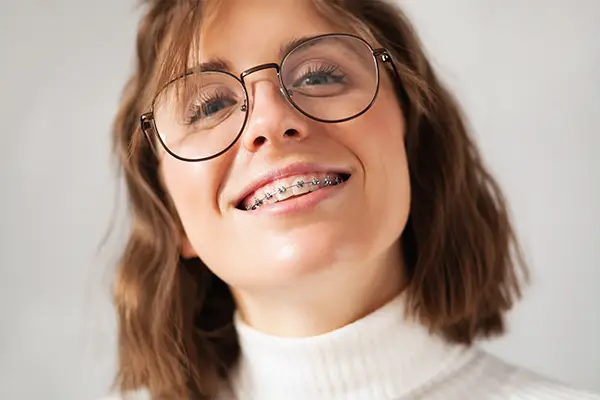 A young woman with glasses smiling brightly, highlighting her traditional metal braces at Riverwind Dental in Richmond, VA